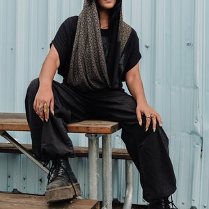 Peut inclure: Un capuchon noir orn&eacute; d'un motif g&eacute;om&eacute;trique d'&eacute;toiles dor&eacute;es r&eacute;p&eacute;titives, port&eacute; sur un t-shirt noir &agrave; manches courtes et un pantalon noir &agrave; jambes larges. La tenue est compl&eacute;t&eacute;e par des bottines noires &agrave; lacets et plusieurs bagues.