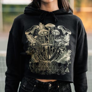 May include: Black cropped hoodie with a gold graphic of mushrooms and a geometric pattern. The hoodie has a drawstring hood.