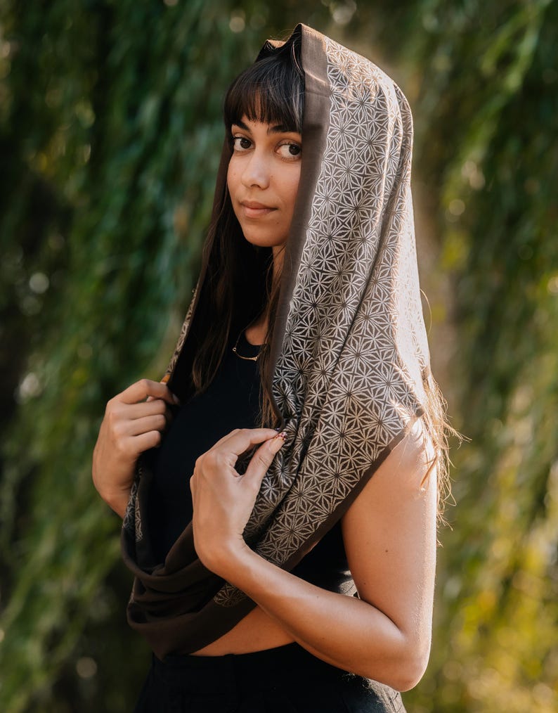 Peut inclure: Une &eacute;charpe infinie marron fonc&eacute; orn&eacute;e d'un motif g&eacute;om&eacute;trique en forme d'&eacute;toile r&eacute;p&eacute;titif dans une teinte beige plus claire. L'&eacute;charpe est drap&eacute;e sur la t&ecirc;te et autour du cou, mettant en valeur une texture de tissu douce et un imprim&eacute; complexe.