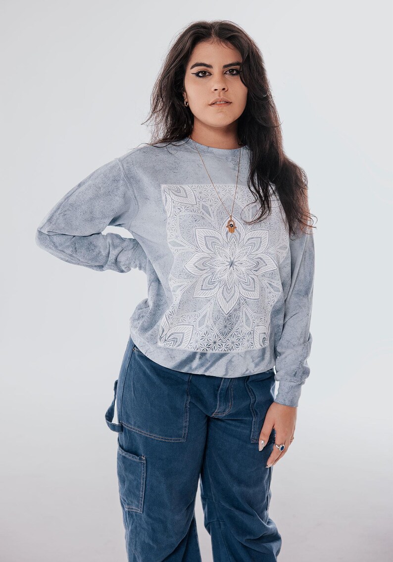 Pode incluir: Uma camisola cinza claro com um desenho de mandala branco. A parte superior de manga comprida &eacute; combinada com cal&ccedil;as de carpinteiro de ganga azul. Um colar de ouro com um pingente &eacute; vis&iacute;vel.