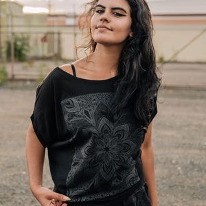 May include: Black off-the-shoulder t-shirt with a large, detailed floral mandala design on the front. The shirt has short sleeves and a relaxed fit. The model is wearing black pants and rings.