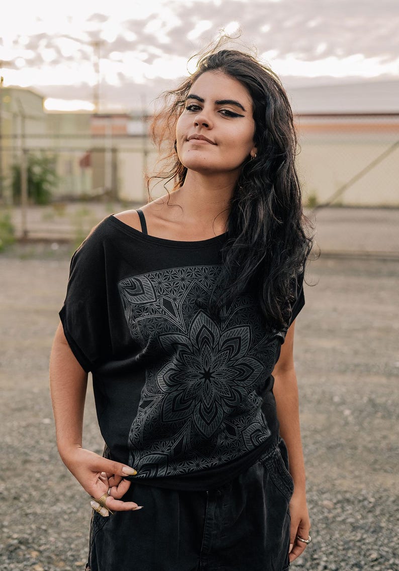 May include: Black off-the-shoulder t-shirt with a large, detailed floral mandala design on the front. The shirt has short sleeves and a relaxed fit. The model is wearing black pants and rings.
