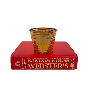 May include: A small, metallic gold-toned bucket with a hammered texture sits atop a bright red hardcover book titled Random House Webster's College Dictionary. The bucket has a tapered shape with a smooth rim and a reflective, polished finish.