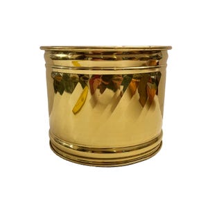 May include: A cylindrical brass planter with a shiny, reflective surface. The planter features a geometric, diamond-shaped pattern around the middle and a rounded rim at the top and bottom. The overall design is simple yet elegant.
