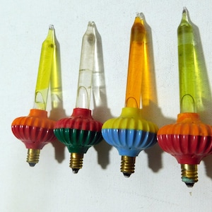 Może przedstawiać: Cztery zabytkowe żarówki choinkowe z kolorowymi podstawami i przezroczystymi, spiczastymi końcówkami. Podstawy są czerwone, pomarańczowe, zielone i niebieskie, z żółtymi, przezroczystymi, pomarańczowymi i zielonymi końcówkami. Żarówki mają klasyczny, retro design.