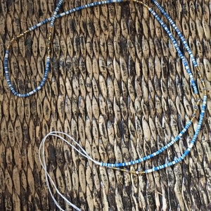 May include: A blue and white beaded necklace with gold accents. The necklace is lying on a woven surface.