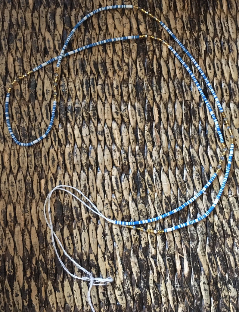 May include: A blue and white beaded necklace with gold accents. The necklace is lying on a woven surface.