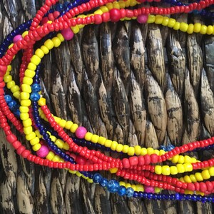 May include: Close-up of colorful beaded necklaces in red, yellow, and blue. The necklaces are made of small, round beads and are arranged in a layered pattern. Some necklaces have pink beads interspersed. The background is a woven texture.