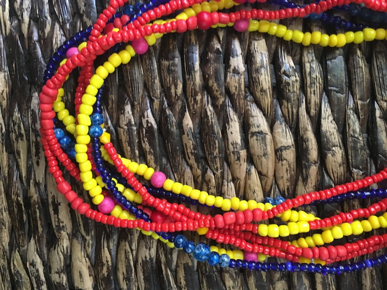 May include: Close-up of colorful beaded necklaces in red, yellow, and blue. The necklaces are made of small, round beads and are arranged in a layered pattern. Some necklaces have pink beads interspersed. The background is a woven texture.