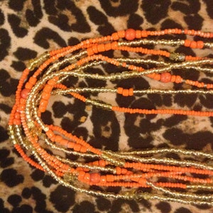 May include: A collection of orange and gold beaded strands. The beads vary in size and shape, with some being small and round, and others being larger and more rectangular. The strands are layered and intertwined, creating a textured and visually appealing design.
