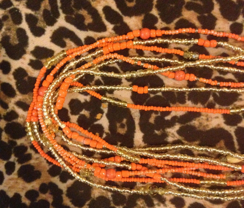 May include: A collection of orange and gold beaded strands. The beads vary in size and shape, with some being small and round, and others being larger and more rectangular. The strands are layered and intertwined, creating a textured and visually appealing design.