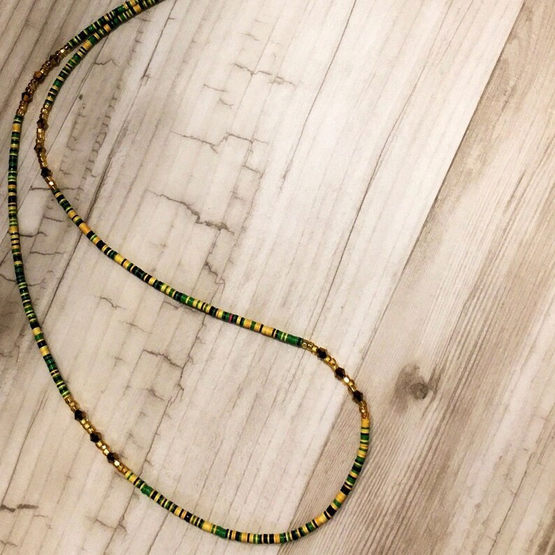 May include: A long, beaded necklace with alternating gold, green, and black beads. The necklace is laid out on a light wood surface.
