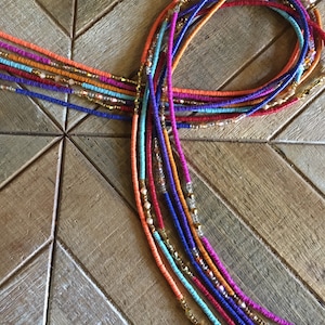 May include: A collection of colorful beaded waist beads in various shades of red, orange, blue, purple, and gold. The beads are arranged on a wooden surface, showcasing their vibrant hues and intricate design. The waist beads are a beautiful accessory.