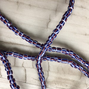 May include: A close-up of a strand of blue, white, and orange striped beads. The beads are strung on a white cord.