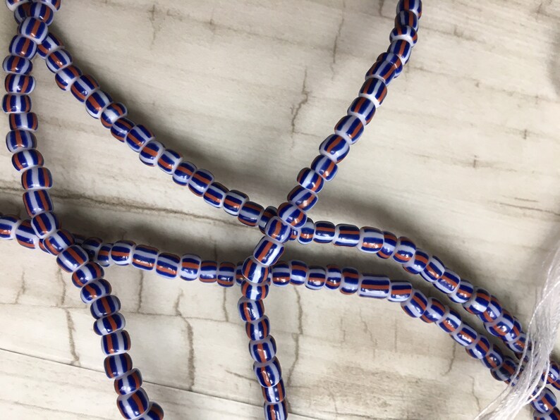 May include: A close-up of a strand of blue, white, and orange striped beads. The beads are strung on a white cord.