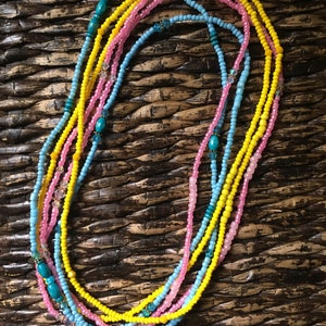 May include: A collection of colorful beaded necklaces in shades of yellow, pink, and blue. The necklaces are arranged in a layered pattern, with turquoise and crystal accents. The beads are small and round, creating a delicate and intricate design.
