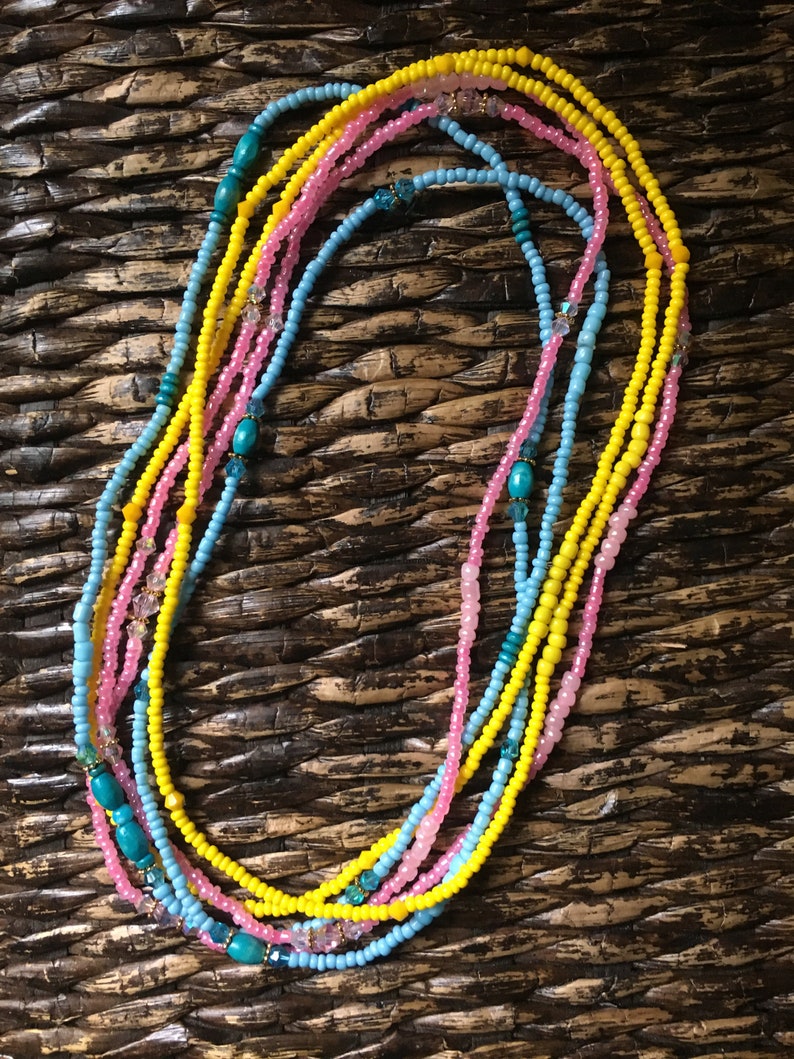 May include: A collection of colorful beaded necklaces in shades of yellow, pink, and blue. The necklaces are arranged in a layered pattern, with turquoise and crystal accents. The beads are small and round, creating a delicate and intricate design.