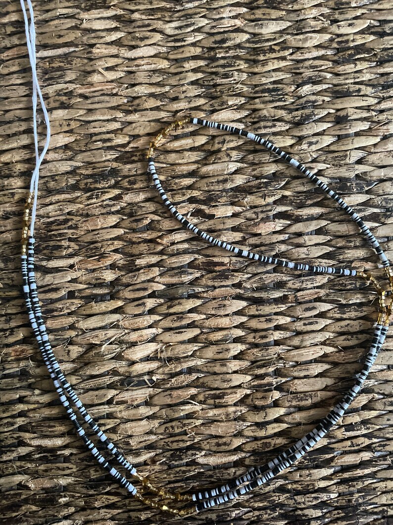 May include: A black and white beaded necklace with gold accents. The necklace is made of small, round beads and is strung on a white cord.
