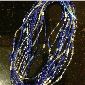 May include: A collection of beaded strands in shades of blue, silver, and white. The beads vary in size and shape, creating a textured appearance. The strands are tied together with a light blue ribbon. The beads are likely used for jewelry or craft projects.