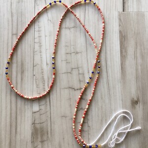 May include: A red and white beaded waist chain with gold and blue accents. The chain is made of small, round beads and has a white cord tie.