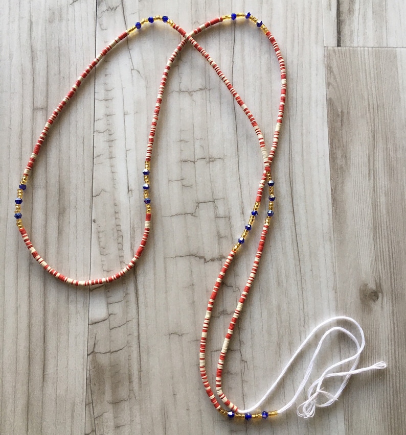 May include: A red and white beaded waist chain with gold and blue accents. The chain is made of small, round beads and has a white cord tie.