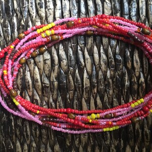 May include: A collection of colorful waist beads. The beads are primarily red, pink, and yellow, with small wooden accents. The beads are arranged in multiple strands, creating a layered effect. The background is a woven, textured surface.