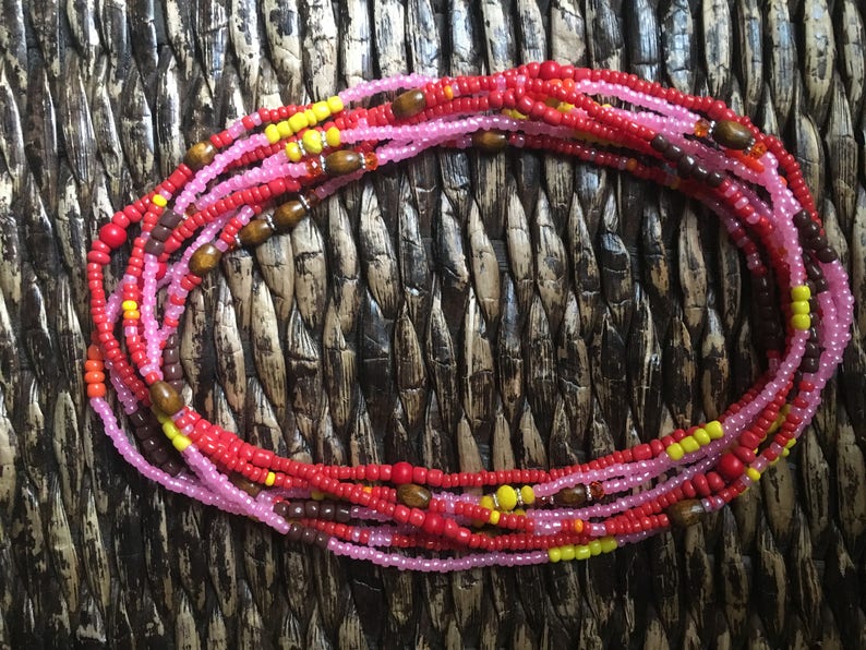 May include: A collection of colorful waist beads. The beads are primarily red, pink, and yellow, with small wooden accents. The beads are arranged in multiple strands, creating a layered effect. The background is a woven, textured surface.