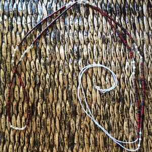 May include: A long, black and red beaded necklace with white beads interspersed throughout. The necklace is lying on a woven surface.