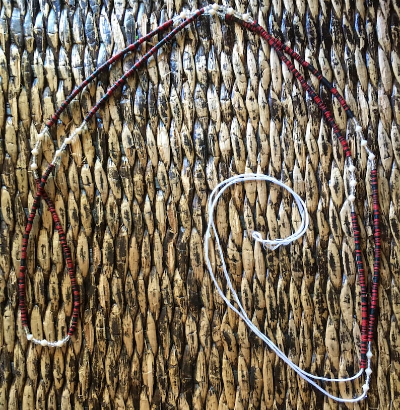 May include: A long, black and red beaded necklace with white beads interspersed throughout. The necklace is lying on a woven surface.