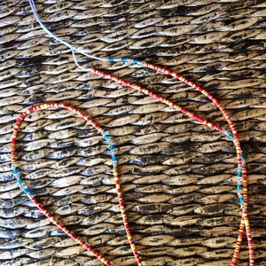 May include: Two beaded necklaces with red, yellow, and blue beads. The necklaces are lying on a woven surface.
