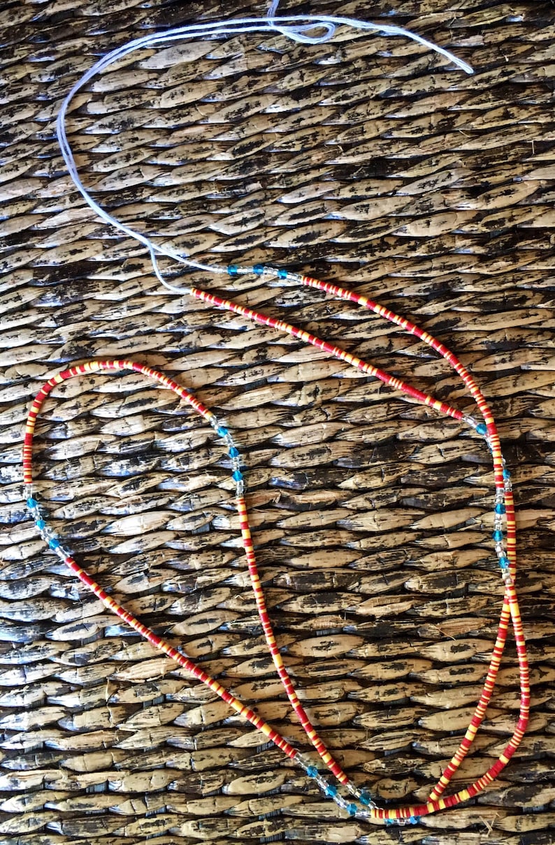 May include: Two beaded necklaces with red, yellow, and blue beads. The necklaces are lying on a woven surface.