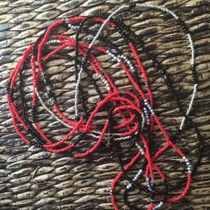 May include: A collection of beaded necklaces in red, black, and silver. The necklaces are made of small, round beads strung together. The beads are arranged in various patterns, creating a layered and textured appearance. The necklaces are displayed on a woven surface.