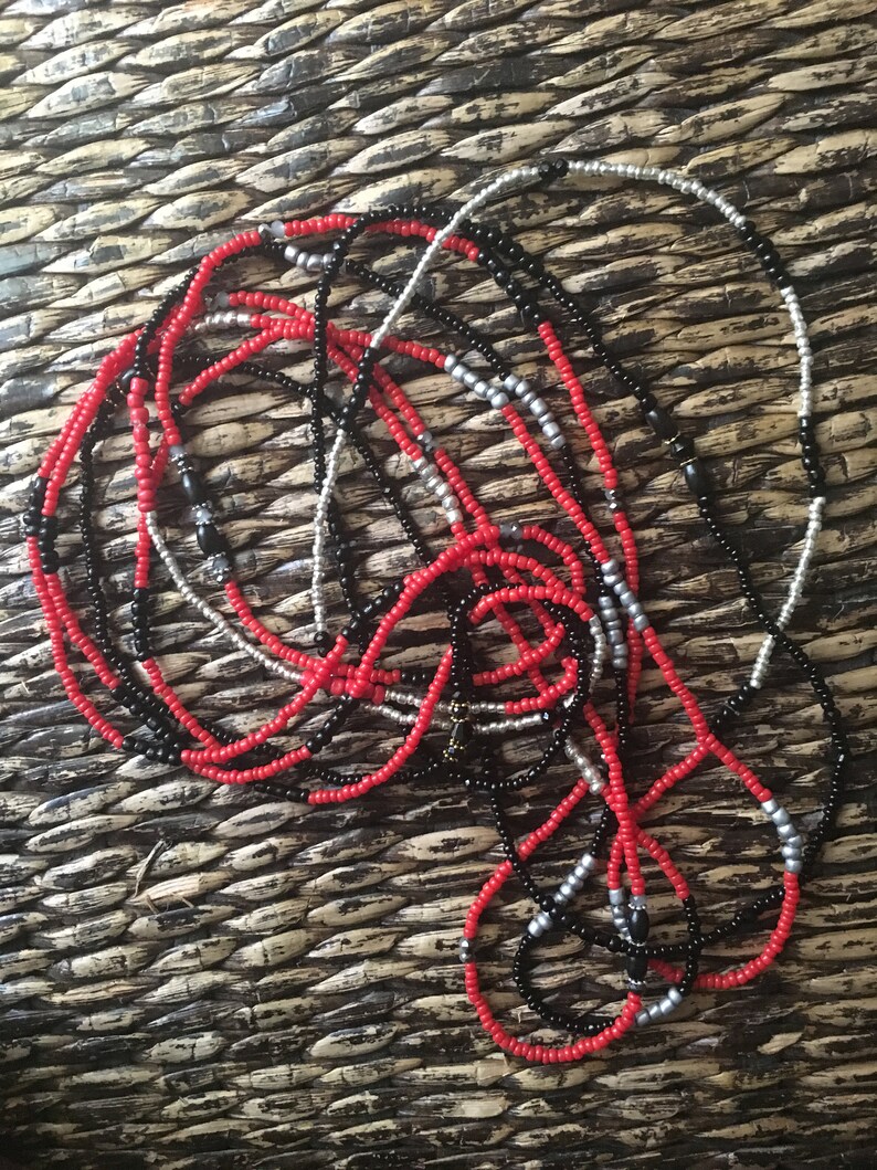 May include: A collection of beaded necklaces in red, black, and silver. The necklaces are made of small, round beads strung together. The beads are arranged in various patterns, creating a layered and textured appearance. The necklaces are displayed on a woven surface.