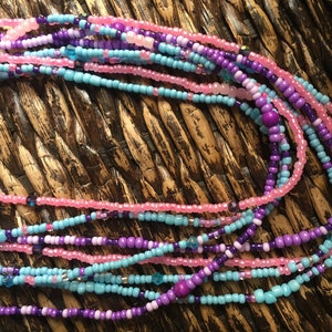 May include: Strands of colorful waist beads in shades of pink, purple, and turquoise. The beads are small and round, with a mix of solid and translucent beads. The beads are strung together, creating a layered effect. The background is a woven texture.