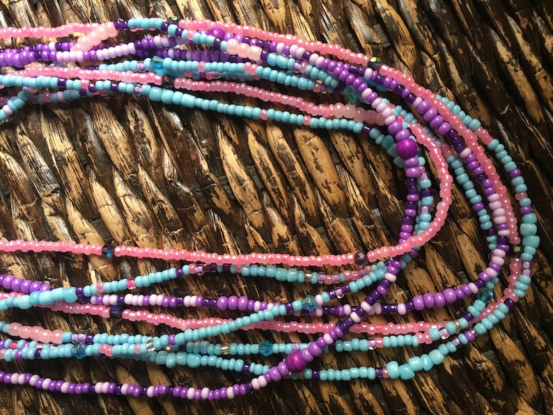 May include: Strands of colorful waist beads in shades of pink, purple, and turquoise. The beads are small and round, with a mix of solid and translucent beads. The beads are strung together, creating a layered effect. The background is a woven texture.