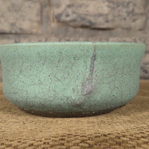 May include: A turquoise ceramic bowl with a crackled texture. The bowl is round with a wide opening. The exterior has subtle grey streaks. The bowl is placed on a brown surface. The bowl is likely handmade.