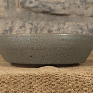 May include: A small, round, gray ceramic bowl with a textured surface. The bowl has a low profile and sits on a slightly raised base. The background is a neutral stone wall.