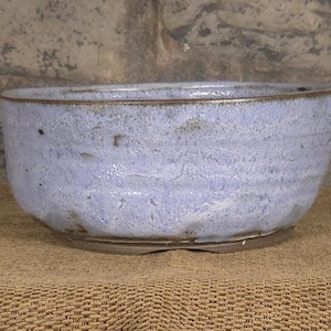 May include: A round ceramic bowl with a textured, light periwinkle blue glaze and a dark brown rim. The exterior surface features subtle horizontal ridges and a mottled finish, while the base is a contrasting dark brown, unglazed clay material.