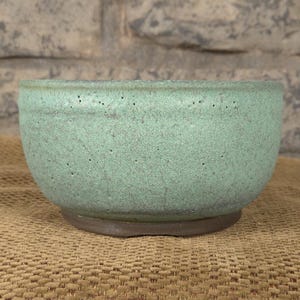 May include: A round, light green ceramic bowl with a textured surface and a dark brown base. The bowl is set against a neutral background, showcasing its simple, yet elegant design. This bowl is suitable for serving food or as a decorative piece.