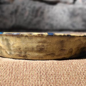 May include: A handmade, rustic ceramic bowl with a textured surface. The bowl features a blend of tan and brown hues, with a subtle blue accent along the rim. The bowl is displayed on a textured, brown surface.