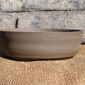 May include: A brown ceramic planter with a smooth, slightly textured finish. The planter is oval shaped and has a wide, shallow opening.