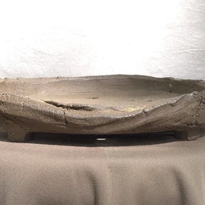 May include: A handcrafted, gray ceramic bonsai pot with a textured, organic design. The shallow, oval-shaped container sits on small feet and features a rough, uneven rim. The neutral color and naturalistic form make it suitable for various plant styles.