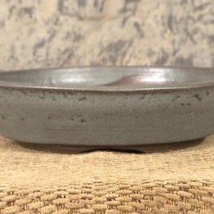 May include: A round, gray ceramic bonsai pot with a textured surface. The pot has a wide, shallow design with a slightly raised rim. The interior has a darker, brownish hue.