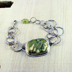May include: A silver bracelet with a green and white stone pendant. The bracelet is made of a chain of small, hammered silver circles.