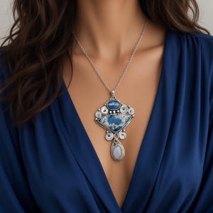 May include: A silver-toned necklace featuring a large, ornate pendant with a triangular blue and white stone, a smaller oval blue stone at the top, and a teardrop-shaped translucent stone dangling at the bottom, all set in intricate metal scrollwork.