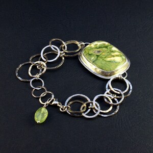 May include: A silver bracelet with a green and white stone pendant and a smaller green stone charm. The bracelet is made of interconnected silver rings.