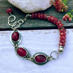 Könnte beinhalten: Ein Armband mit drei ovalen, roten Cabochon-Steinen in Silberfassungen, die durch Silberglieder verbunden sind und an eine Reihe facettierter roter Perlen anschließen. Das Schmuckstück besitzt einen Karabinerverschluss und verstellbare Ringe.