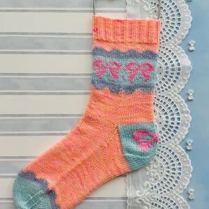 May include: A hand-knitted sock in shades of coral, teal, and pink, featuring a bow pattern. The cuff, heel, and toe are teal, while the main body is coral with a teal and pink bow design. The sock is hanging on a metal hanger.