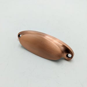 May include: A brushed copper oval cabinet pull with rounded edges and two small holes for mounting.