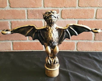 Könnte beinhalten: Eine goldfarbene Metall-Gargoyle mit Fledermausflügeln und einem bedrohlichen Ausdruck. Die Gargoyle sitzt auf einem runden Sockel und hat zwei kleine Kerzenhalter an beiden Seiten.